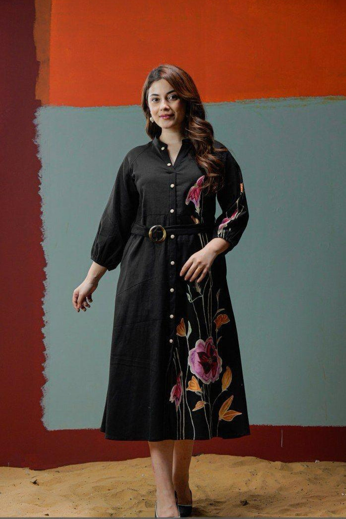 Woman in a black dress with floral embroidery standing against a colorful abstract wall.